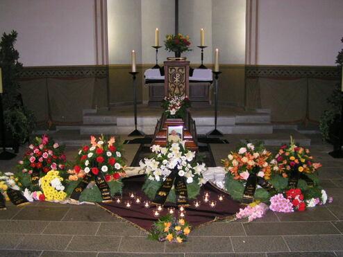 Blumenarrangements in verschiedenen Farben umgeben einen mit Kerzen und einem Altar geschm&uuml;ckten Sarg in einer Kirche.