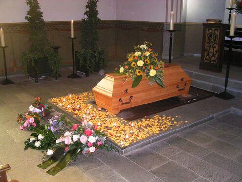 Holzkasten mit Blumen und Bl&uuml;tenbl&auml;ttern, umgeben von Kerzen in einer Kirche.