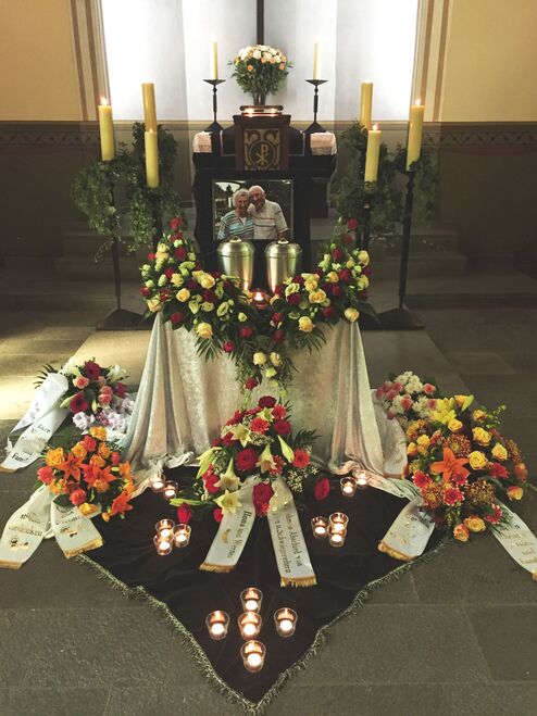 Gedenkst&auml;tte mit Blumenarrangements, Kerzen und einem Bild von zwei Personen sowie Urnen in einem ruhigen Raum.