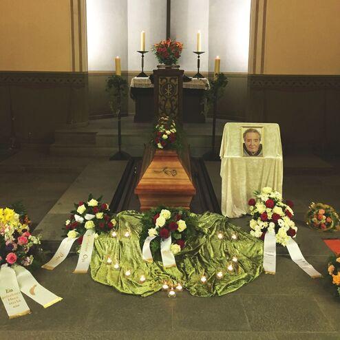 An einem Sarg stehen Blumenarrangements und Kerzen, dahinter ein Portr&auml;t der verstorbenen Person.