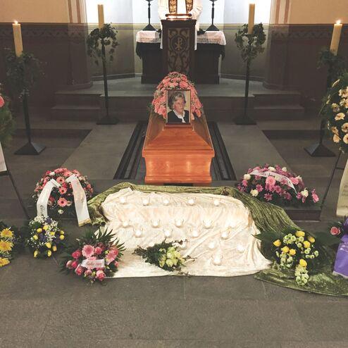 Ein Sarg mit Blumenarrangements und Kerzen im Hintergrund einer Trauerfeier in einer Kirche.
