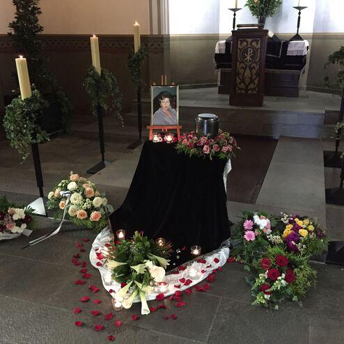 Gedenktafel mit einem Foto, Kerzen und Blumenarrangements in einer Trauerhalle.