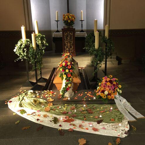 Ein mit Blumen geschm&uuml;ckter Sarg in einer Kapelle, umgeben von Kerzen und dekorativen Pflanzenarrangements.
