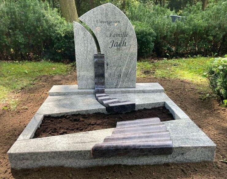 Granitgrabstein mit Inschrift &bdquo;Familie J&auml;sch&ldquo; und stilisierter Treppe in einem ruhigen Landschaftsbild.
