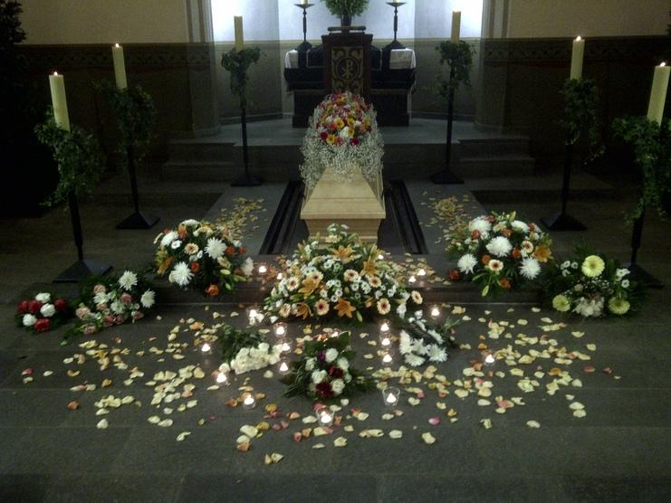 Blumenarrangements und Kerzen umgeben einen ged&auml;mpften Sarg auf einem altar&auml;hnlichen Podest.