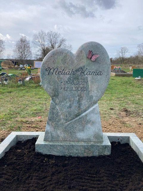 Herzf&ouml;rmiger Grabstein mit der Inschrift &bdquo;Meliah Kama&ldquo; und einem Schmetterlingsmotiv, umgeben von unbepflanztem Erdreich.