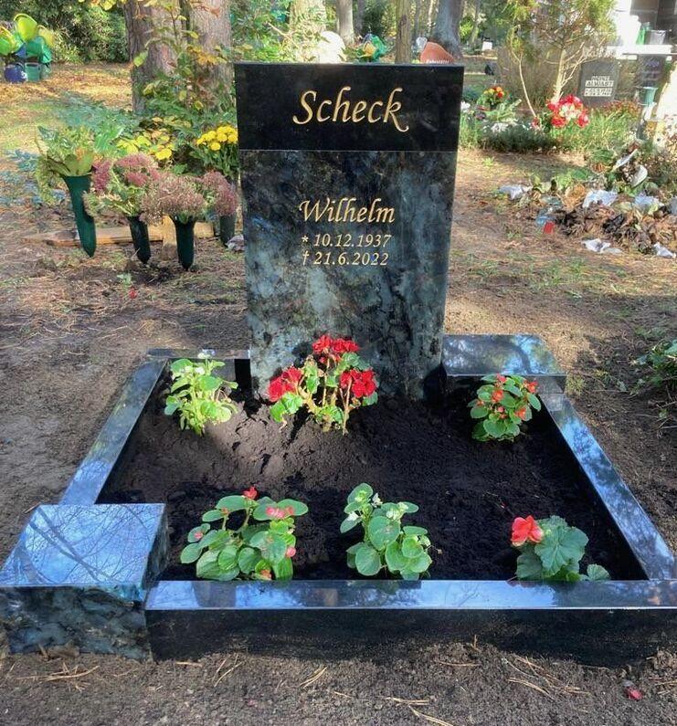 Schwarzer Grabstein mit der Inschrift "Scheck" und "Wilhelm", umgeben von roten Begonien auf schwarzem Erdgrund.