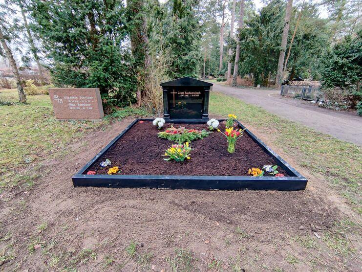Schwarzes Grab mit Blumen und Licht auf einem Friedhof, umgeben von B&auml;umen und Gedenktafel im Hintergrund.