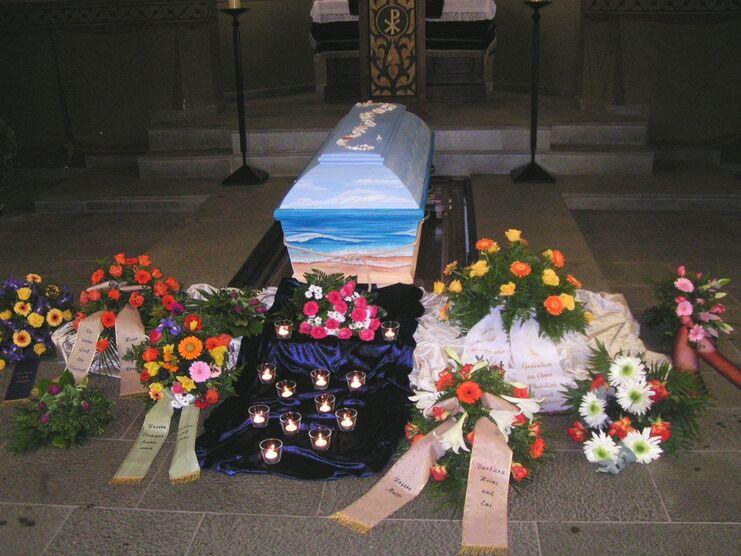 Blick auf einen bunten Sarg, umgeben von Blumenarrangements und Kerzen in einem Kirchenraum.
