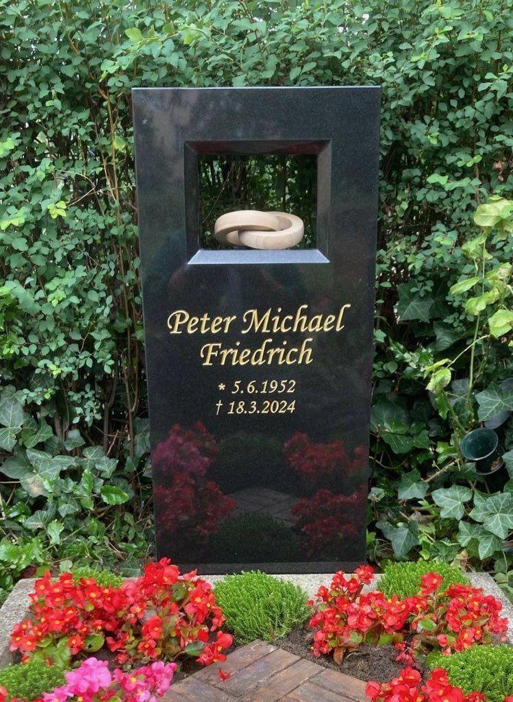 Schwarzer Grabstein mit Inschrift f&uuml;r Peter Michael Friedrich, umgeben von bunten Blumen und gr&uuml;ner Bepflanzung.