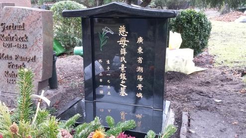 Schwarzes Grabmal mit goldenen Schriftzeichen und floralen Motiven, umgeben von Gr&uuml;npflanzen und dekorativen Elementen.