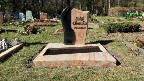 Grabstein aus hellem und dunklem Stein auf einem Friedhof, umgeben von Blumen und weiterer Grabdekoration.
