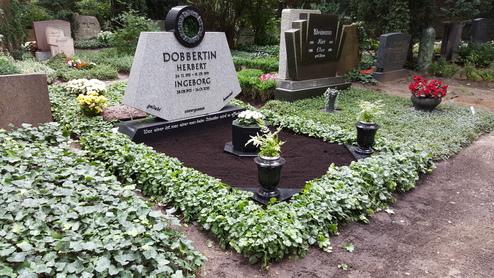 Grabh&uuml;gel mit grauem Steingrabstein, umgeben von Pflanzen und Blumen auf einem ruhigen Friedhof.