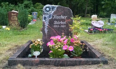 Grabstein aus schwarzem Granit mit Sonnenblumenmotiv, umgeben von bunten Blumen auf einer Friedhofsanlage.