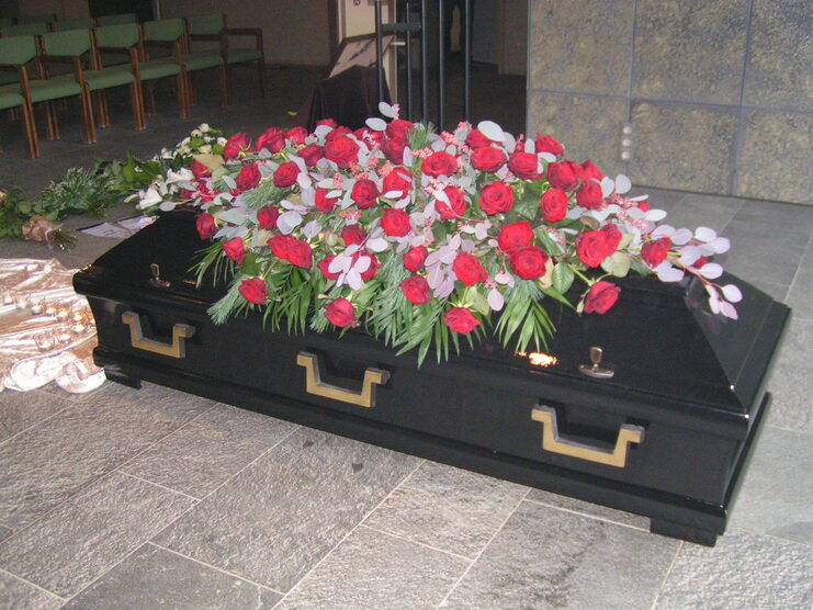 Schwarzer Sarg mit roten Rosen und gr&uuml;nem Laub als Dekoration, auf einem schlichten Untergrund in einem Raum.