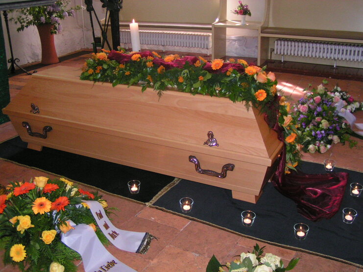 Holzleichnam mit Blumen und Kerzen, umgeben von Trauergestecken in einem hellen Raum.