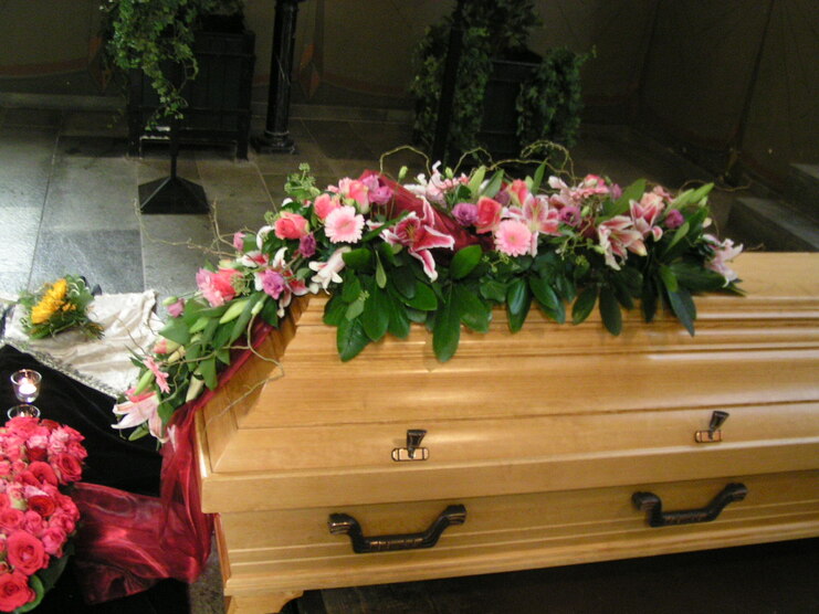 Holzkahn mit bunten Blumenarrangements und gr&uuml;nen Bl&auml;ttern, umgeben von dunklem Raum und Trauerschmuck.