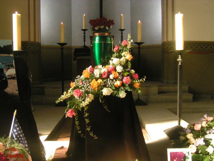 Gr&uuml;ner Urnenbeh&auml;lter mit bunten Blumen auf schwarzem Tuch in einer lichtdurchfluteten Kapelle, umgeben von Kerzen.
