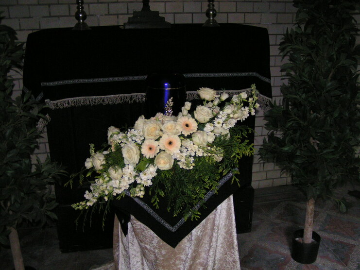 Blumenarrangement aus wei&szlig;en Rosen und anderen Blumen auf einem schwarzen Tuch vor einer Urne in einem Trauerraum.