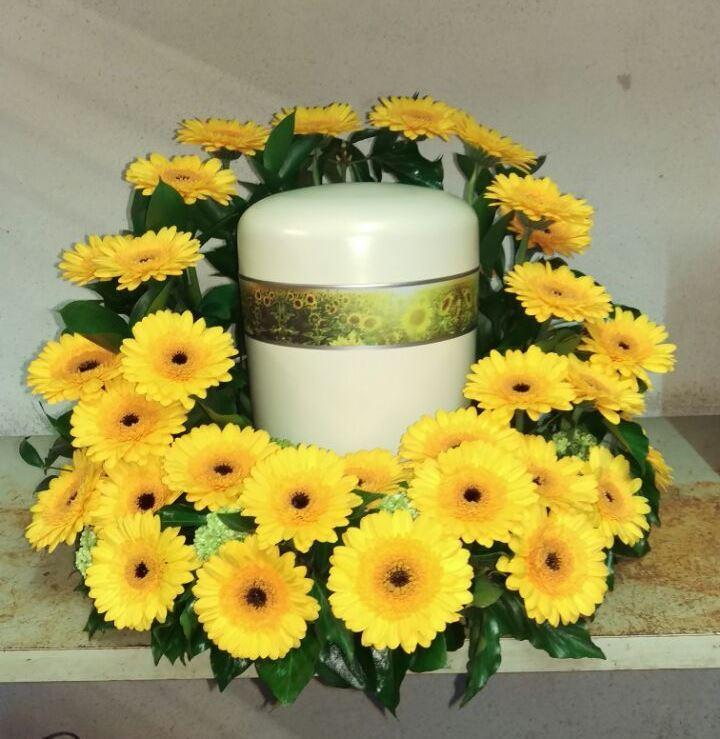 Wei&szlig;er Urnenbeh&auml;lter umgeben von gelben Sonnenblumen und gr&uuml;nen Bl&auml;ttern auf einer neutralen Oberfl&auml;che.