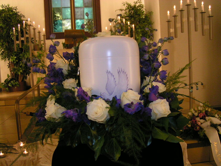 Wei&szlig;er Urnensarg umgeben von lila und wei&szlig;en Blumen in einer feierlichen Trauerumgebung.