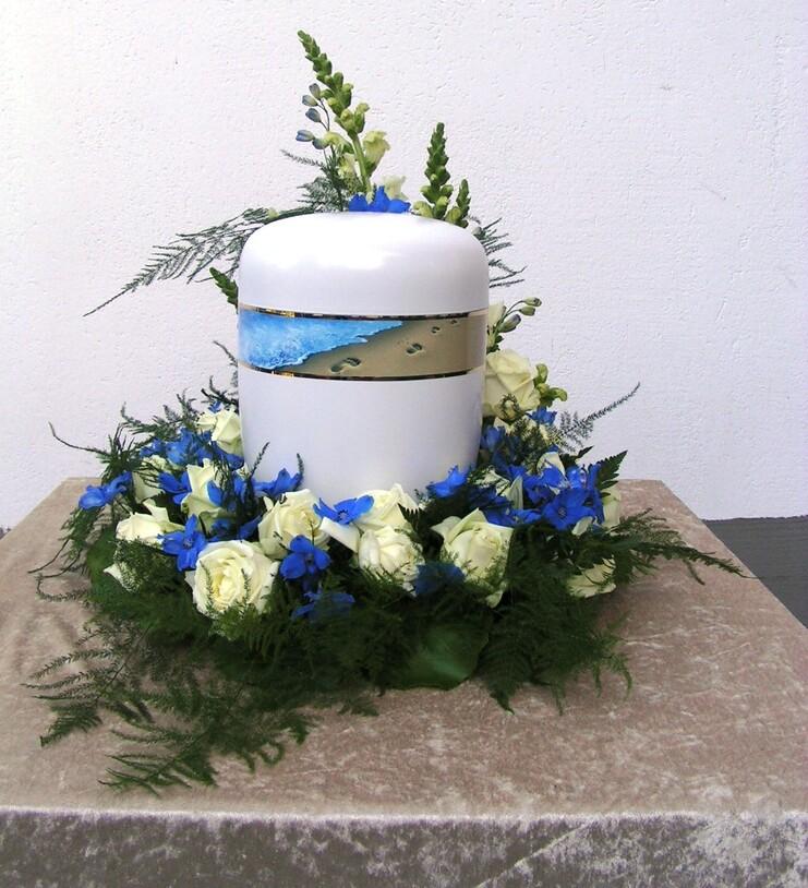 Wei&szlig;e Urne verziert mit blauen und wei&szlig;en Blumen, umgeben von gr&uuml;nen Bl&auml;ttern auf einem beigen Tisch.