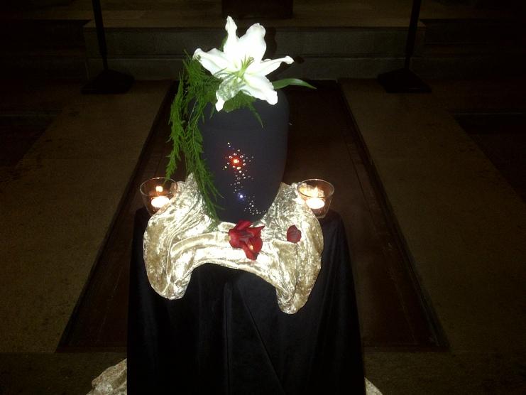 Schwarze Urne mit wei&szlig;en Lilien und gr&uuml;ner Dekoration auf einem edlen Tuch, umgeben von Kerzen und Rosenbl&uuml;ten.