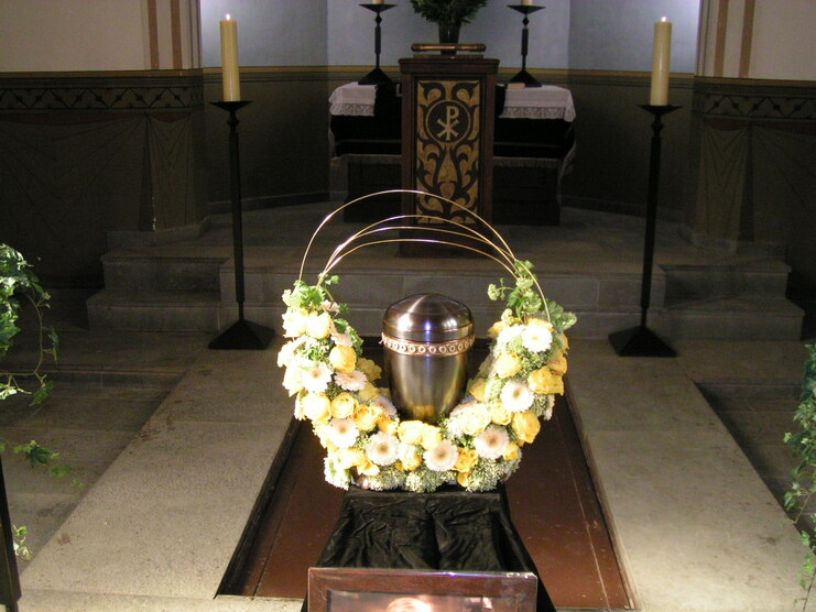 Urne mit Blumenkranz auf einem Tisch in einer Kirche, umgeben von Kerzen und einem Altar im Hintergrund.