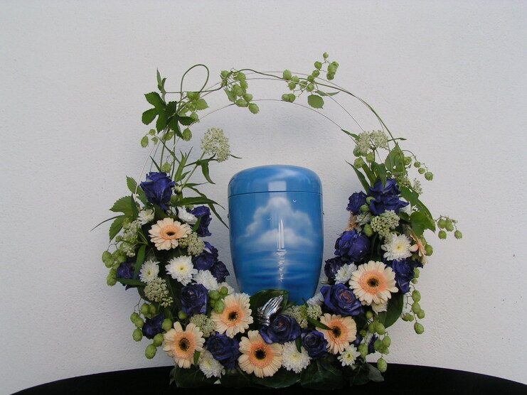 Blaues Urnengedeck umgeben von wei&szlig;en und lila Blumen in einem floralen Kranz auf einem schwarzen Untergrund.
