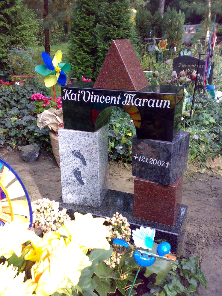 Grabstein aus mehreren Steinbl&ouml;cken mit Inschrift &bdquo;Kai Vincent Maraun&ldquo; und Fu&szlig;abdr&uuml;cken, umgeben von Blumen und Dekorationen.