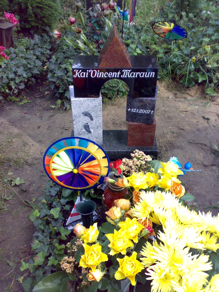 Grabstein mit der Inschrift &bdquo;Kai Vincent Maraun&ldquo; und bunten Blumen sowie einer farbigen Windm&uuml;hle im Vordergrund.