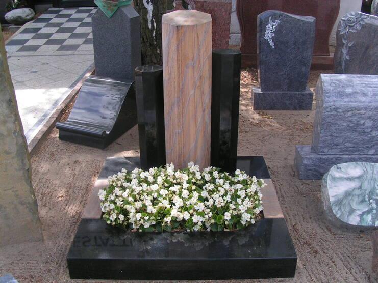 Grabanlage mit einem hohen, gestreiften Steins&auml;ulen-Mittelst&uuml;ck und wei&szlig;en Blumen auf schwarzem Untergrund.