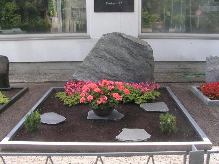 Grabst&auml;tte mit einem gro&szlig;en Stein, bunten Blumenbeeten und grauen Platten in einem gepflegten Bereich.