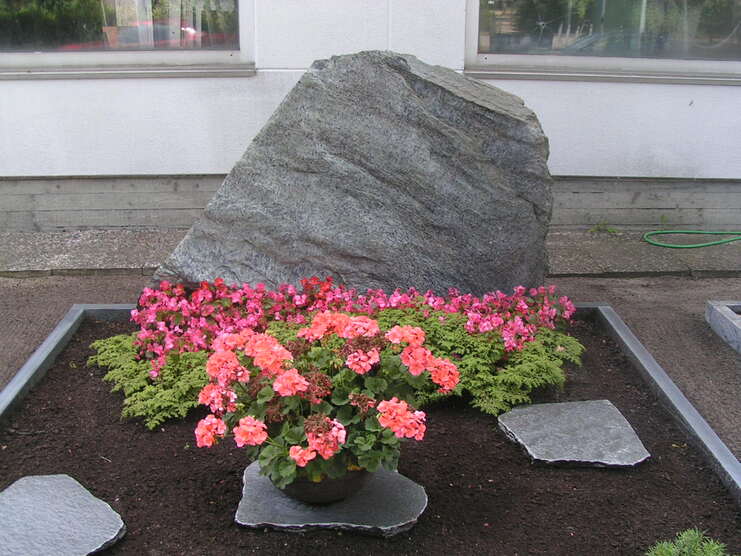 Graniterner Stein hinter bunten Begonien auf einem bepflanzten Beet vor einem Gebäude.