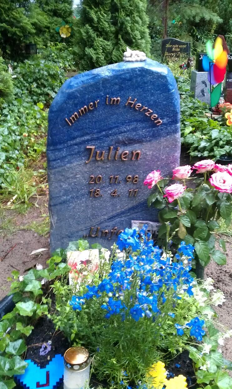 Grabstein mit Inschrift &bdquo;Immer im Herzen, Julien, 20.11.98 &ndash; 18.4.17&ldquo;, umgeben von bunten Blumen und Dekorationen.
