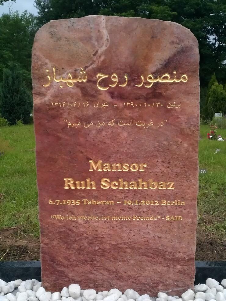Grabstein aus r&ouml;tlichem Stein mit Inschriften zu Mansor Ruh Schahbaz, Jahr und Zitat auf einem Kiesbett.