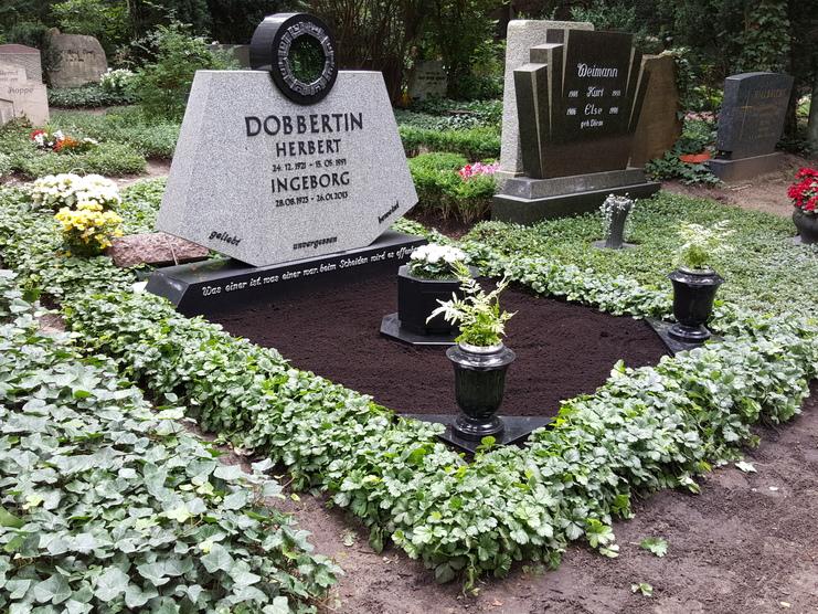Grabstein mit Inschrift „DOBBERTIN Herbert und Ingeborg“ umgeben von Pflanzen und Blumen auf einem ruhigen Friedhof.
