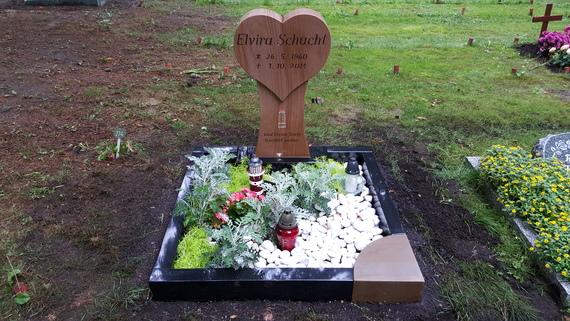 Grabanlage mit herzf&ouml;rmigem Holzstele und Blumen, umgeben von wei&szlig;en Steinen und roten Kerzen auf gr&uuml;nem Rasen.