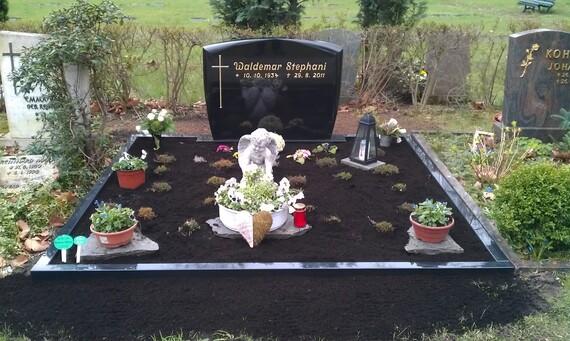 Grabst&auml;tte mit schwarzem Grabstein, Blumenschmuck, einer Statue und Grablicht auf frischer Erde im Friedhof.