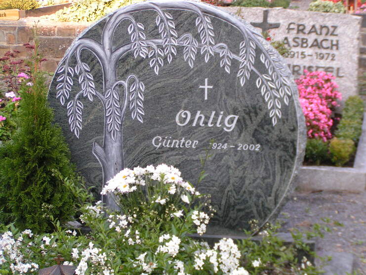 Granitgrabstein mit Baum-Motiv, Beschriftung "Ohlig G&uuml;nter 1924-2002", umgeben von bl&uuml;henden Blumen.