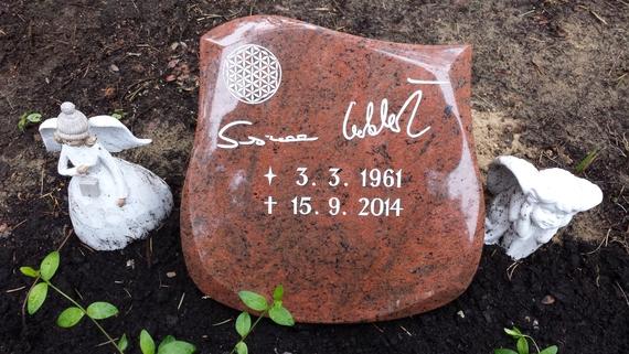 Grabstein aus rotem Granit mit Inschriften und zwei Engel-Statuen auf einer blumenbewachsenen Erde.