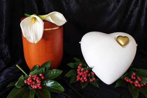 Braune Urne mit Calla-Blumen und eine weiße Herzurne mit goldenen Details, umgeben von grünen Blättern und roten Beeren.