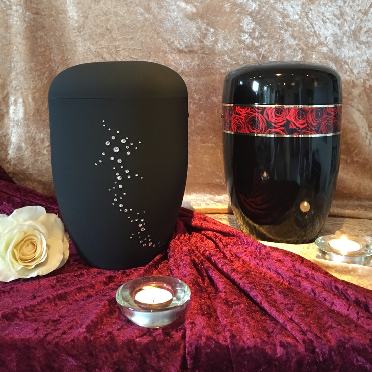Zwei dekorative Urnen auf Samt, eine schwarze mit floralem Muster, die andere gl&auml;nzend mit roten Rosenband, umgeben von Kerzen.