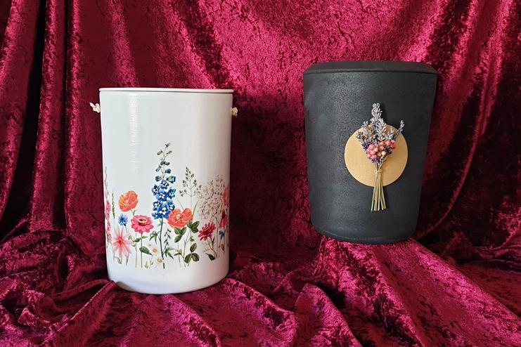 Zwei dekorative Behälter: Ein weißer mit Blumenmuster und ein schwarzer mit floralem Anhänger auf rotem Samt-Hintergrund.