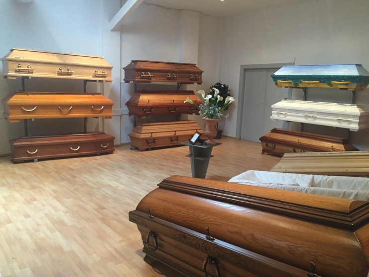 Ausstellung von Holzs&auml;rgen in einem Raum, mit f&uuml;nf S&auml;rgen an W&auml;nden und einem wei&szlig;en Sarg im Vordergrund.