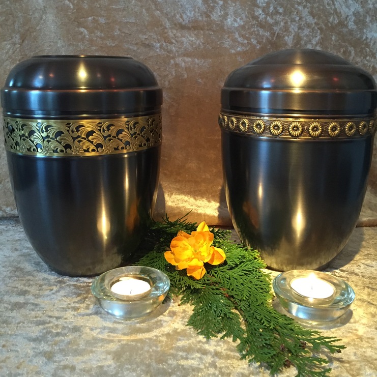 Zwei elegante Urnen mit goldenen Verzierungen, umgeben von Kerzen und einer orangefarbenen Blume auf einem gedeckten Tisch.