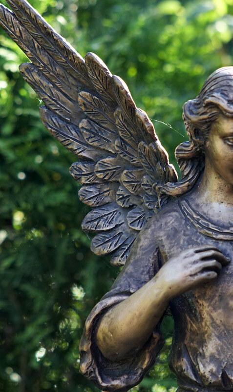 Bronzene Engelstatue mit detaillierten Fl&uuml;geln, die im Gr&uuml;nen steht, mit einem sanften Ausdruck und erhobenem Arm.