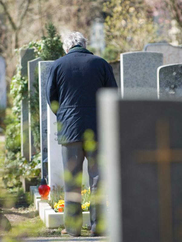 Ein Mann in einem blauen Mantel steht vor einem Grab mit Blumen und einer roten Laterne auf einem Friedhof.