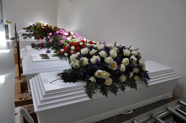Serie von weißen Särgen, geschmückt mit bunten Blumenarrangements, in einem hell erleuchteten Raum.