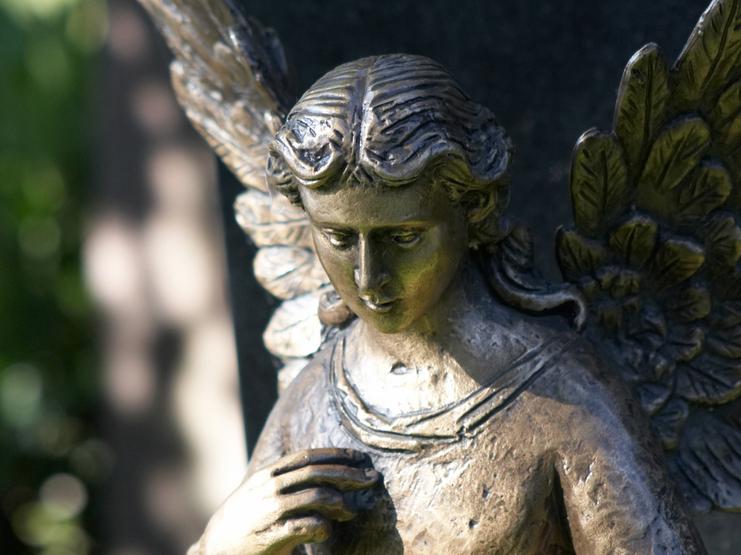 Bronzestatue eines Engels mit gefalteten Händen, stehend vor einem grauen Grabstein im Hintergrund.